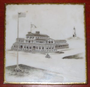Ceramic tile with image of a large hotel and American flag at center. Monument in back.