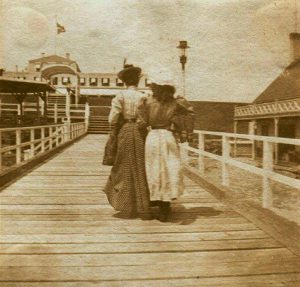 Two women in antique dresses with their backs to camera, on wooden walk, hotel in background.